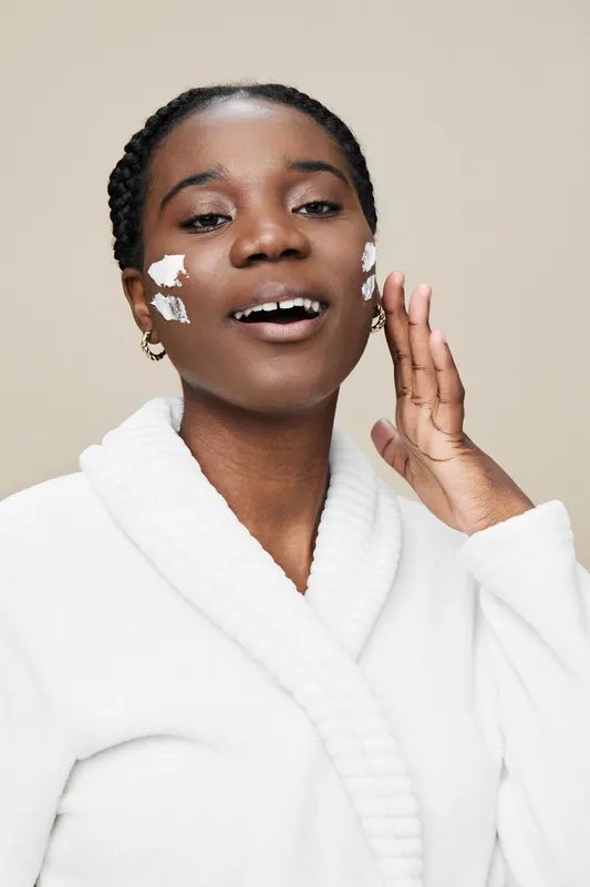 Young African woman applying skincare cream on her face, smiling joyfully in a white robe, showcasing beauty and self care concepts in a neutral background