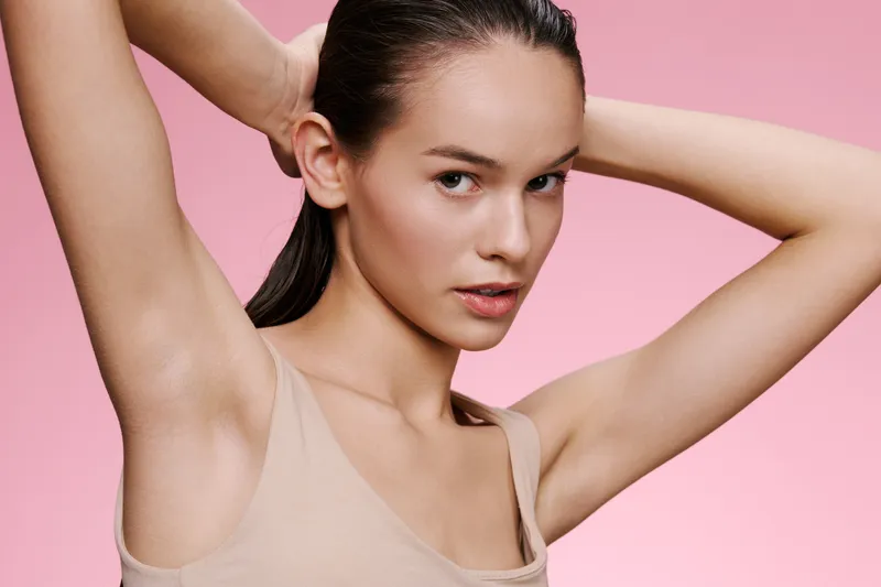 A young female model with long hair poses elegantly with her arms raised, showcasing smooth skin against a soft pink background, emphasizing beauty and confidence