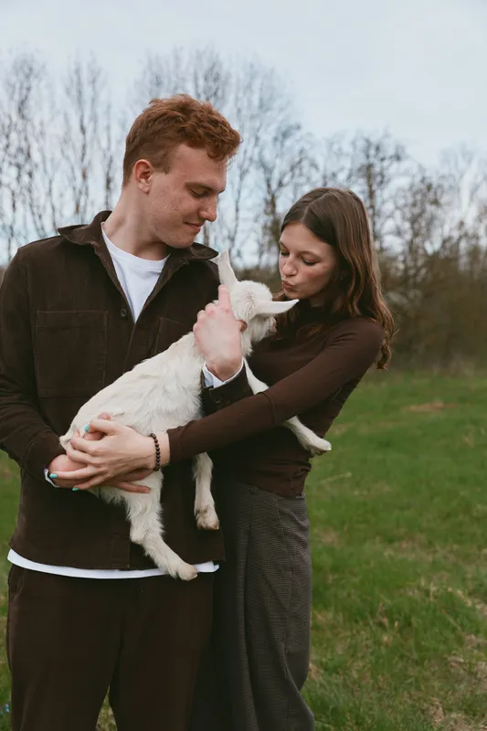 Young couple holds a white baby goat together outdoors in early spring while sharing a tender moment in casual warm clothing on a grassy field with bare trees in the background.