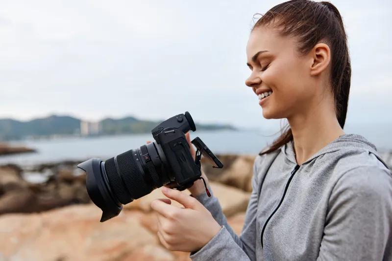 Young woman smiling while holding a professional camera, wearing a grey hoodie, at a coastal location with a blurred sea background
