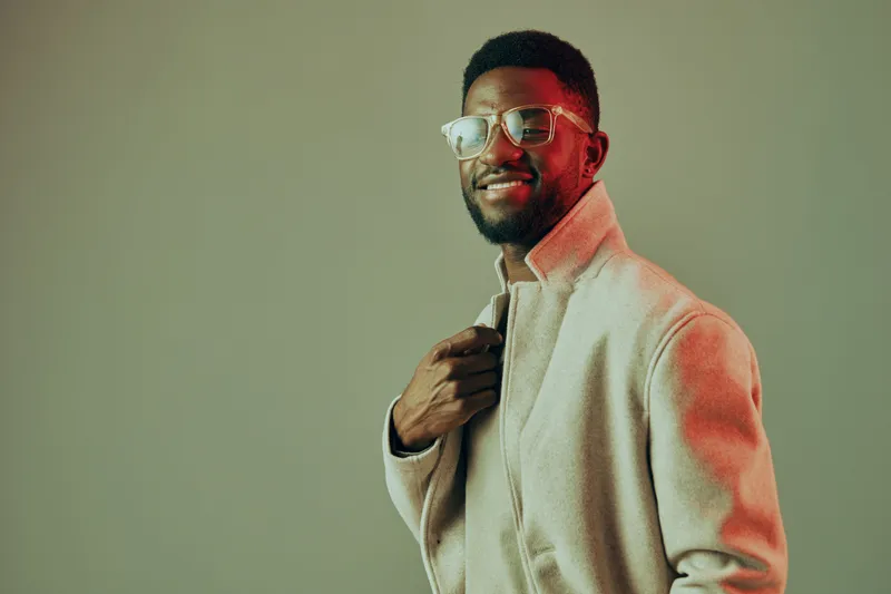 Stylish man in a beige coat smiling, wearing glasses with a soft colorful background, reflecting confidence and modern fashion trends