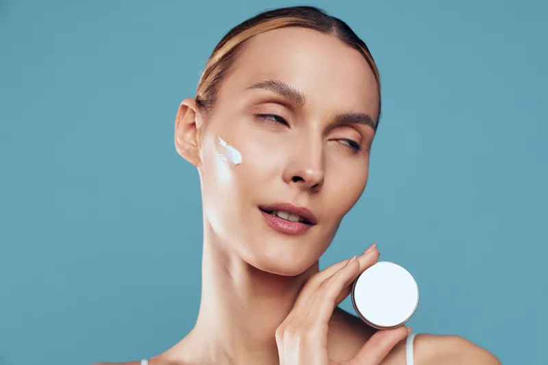Young woman with light skin applies white face cream on her cheek in a close-up against a soft blue background, conveying skincare, wellness, and self care.