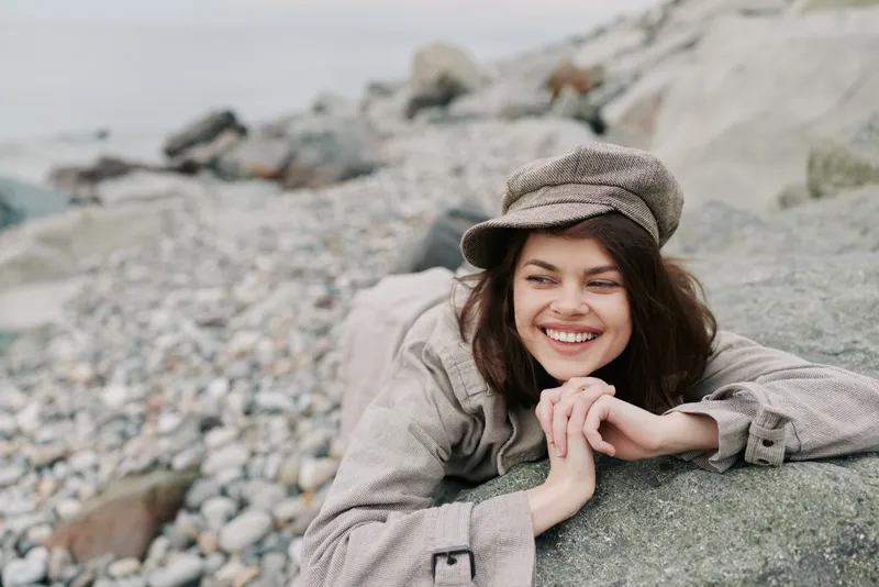 Smiling young woman in casual clothes and hat lies on rocky beach, looking away with relaxed expression. Concept of leisure, nature, and happiness outdoors by the sea.