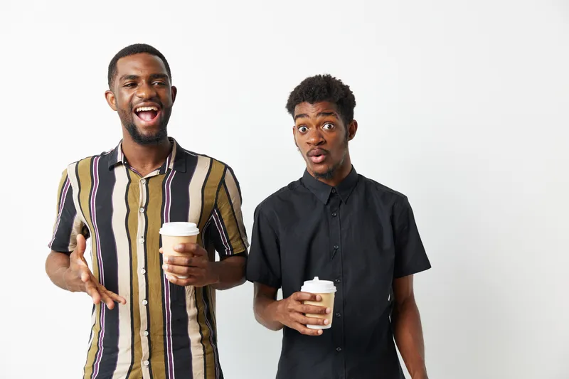 Two smiling African American men enjoying coffee together, dressed casually with vibrant patterns in a bright, minimalist space. A moment of joy captured in everyday life.