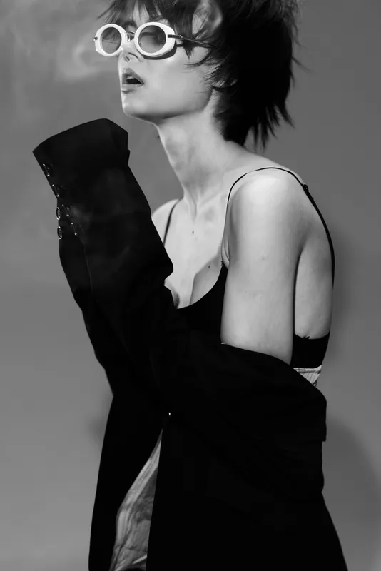 Black and white portrait of a stylish young woman wearing sunglasses and an off-shoulder jacket, expressing confidence and elegance.
