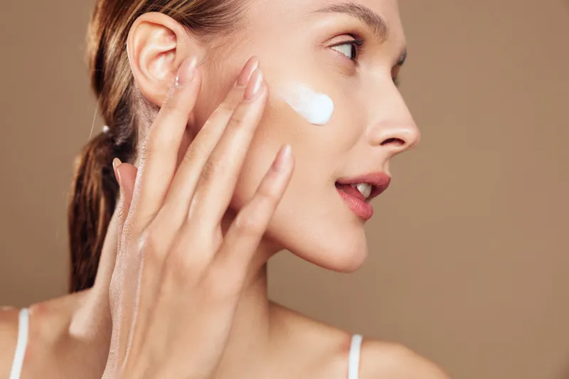 Young woman applying facial cream with fingers, side profile view, clear skin, natural makeup, soft lighting, neutral background, skincare routine concept.