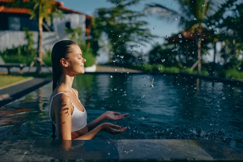 A relaxed woman in a white swimsuit sits at the edge of a sunny pool, water droplets glistening in the air as she smiles and plays with the spray. A lifestyle moment of leisure and wellness.