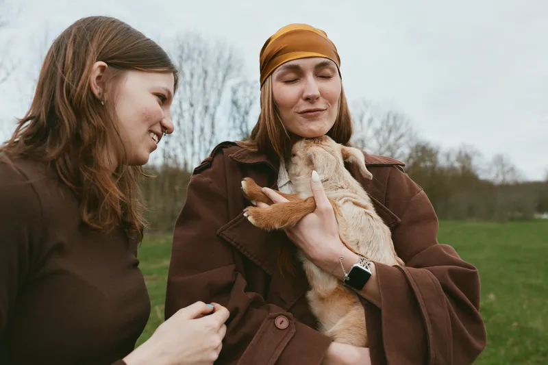 Cheerful woman outdoors with a playful goat kid, smiling and relaxed, sunlit field scene, rustic outfits, gentle breeze, natural colors, countryside backdrop, moment of friendship