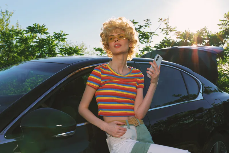 Young woman with curly blond hair stands beside a dark car on a sunny day, wearing a striped crop top and light shorts, posing confidently with a handheld phone