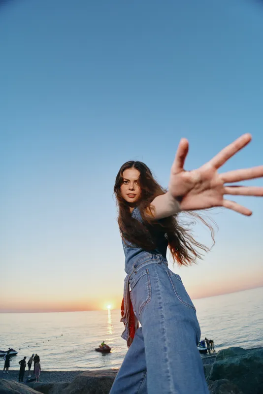 lifestyle woman in modern boho-western denim street style reaches out with hand, film color, casual sunset ocean background, relaxed atmosphere