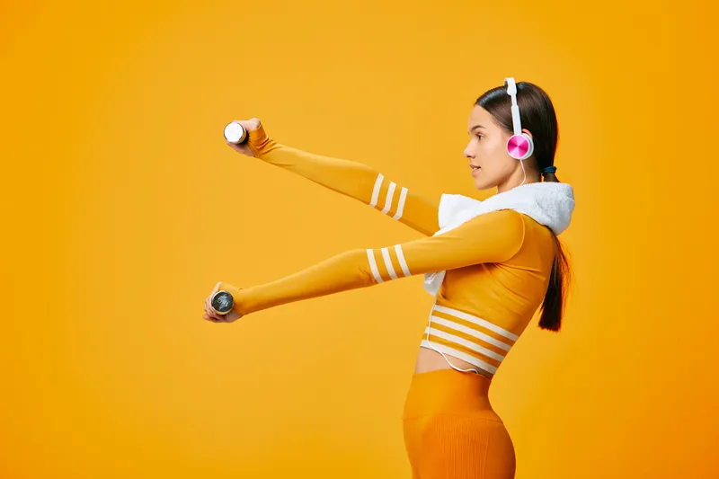 Woman fitness athlete stretches arms with dumbbells in a bright orange studio, wearing headphones and a sporty outfit. Energetic training moment focused on strength and movement.