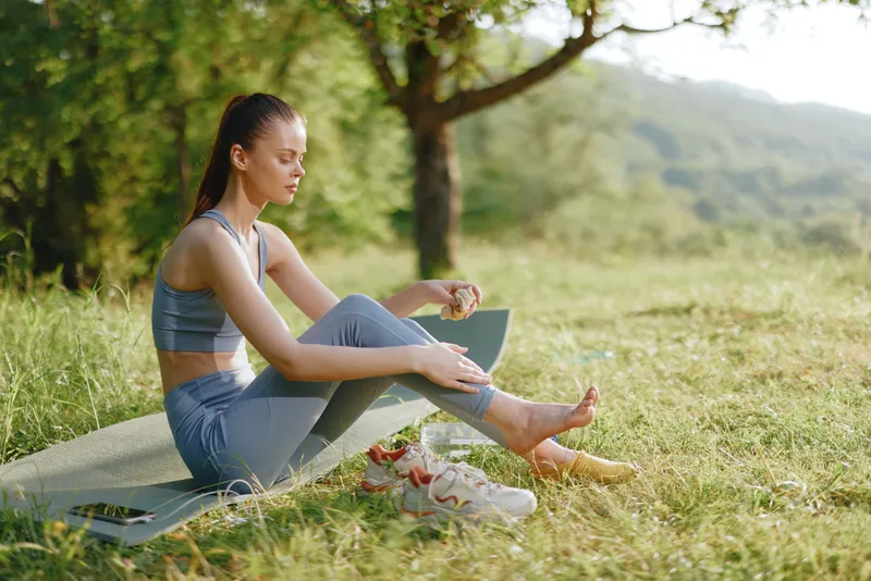 Young woman practicing yoga on a mat outdoors, wearing workout clothes, enjoying healthy lifestyle in nature, with greenery and bright sunlight in the background