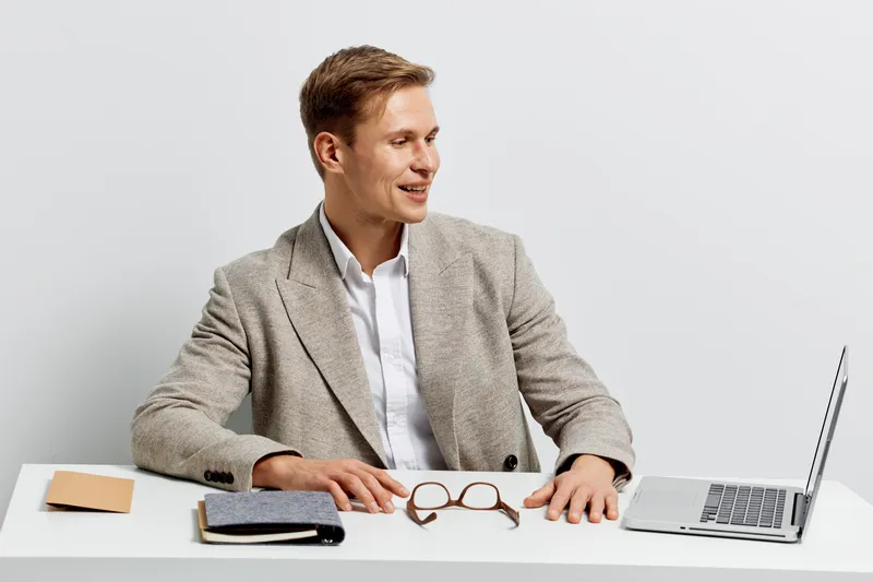 Smiling man in beige blazer sitting at desk with laptop and glasses in bright modern office environment, looking away from the camera. Business casual, professional appearance, concept of work, commun