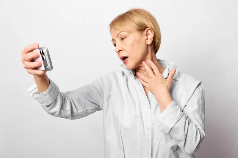 Woman holding smartphone for selfie, hand on neck, pain and surprised expression, senior portrait, white background, health stress and discomfort, soft lighting