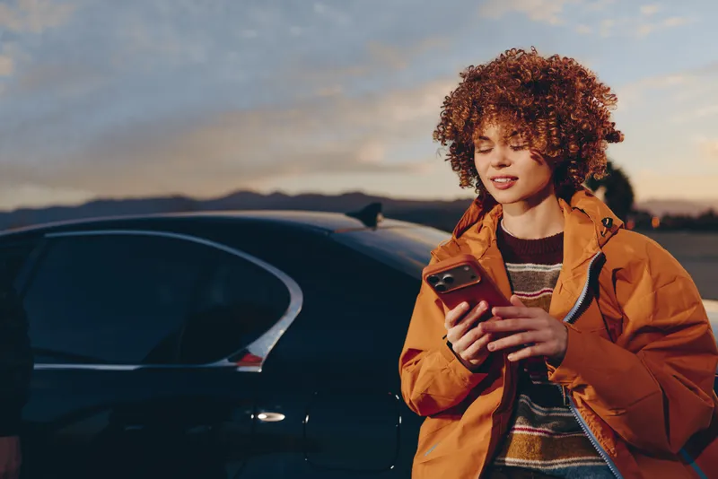 Woman in orange jacket holds a smartphone beside a parked car at sunset, curly hair framing her face, outdoor lifestyle scene with mountains in the distance, calm communication in warm light