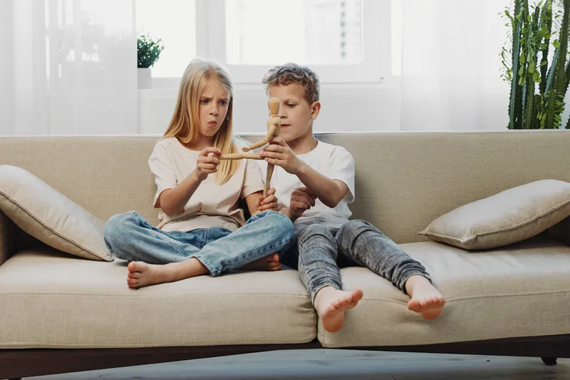 Two children sitting on beige sofa playing with wooden airplane toy. Casual home interior with bright natural light. Concept of childhood, creativity, and shared playtime in family environment.
