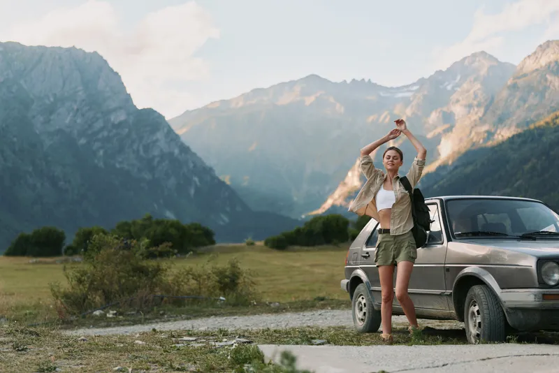 Young woman lifestyle standing near car in mountain landscape, casual outfit, relaxed pose, sunny day, natural scenery, adventure travel concept