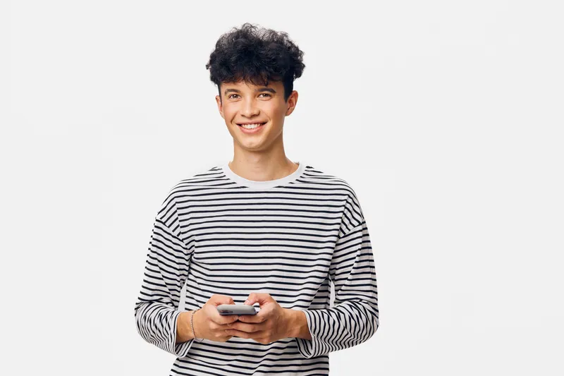 young man smiling holding smartphone, casual striped shirt, curly hair, standing on white background, modern and happy expression in bright lighting