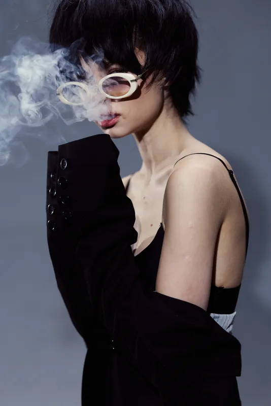 Smoking woman with short hair wearing glasses and black jacket emitting smoke against gray background.
