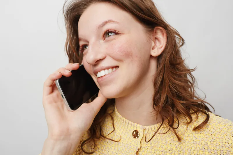 Smiling young woman with curly hair wearing yellow sweater talks on smartphone. Close-up portrait on white background. Concept of communication, happiness, and casual lifestyle.