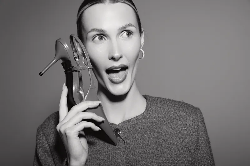 Young white woman holds high heel shoe near face in surprised expression, wearing sweater and hoop earrings on plain background. Concept for fashion, style, and footwear.