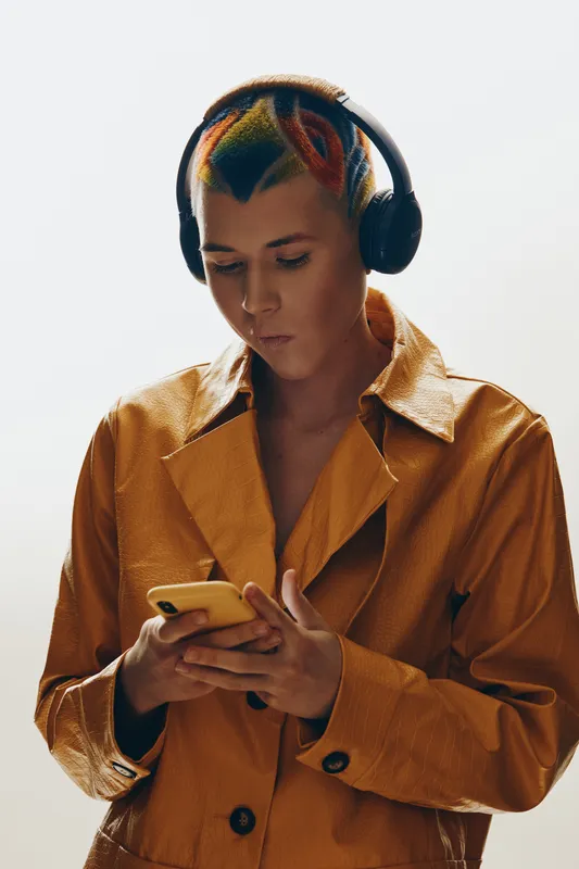 Young woman with colorful dyed hair wearing headphones and a brown jacket focused on her smartphone, isolated on white background, modern urban style.