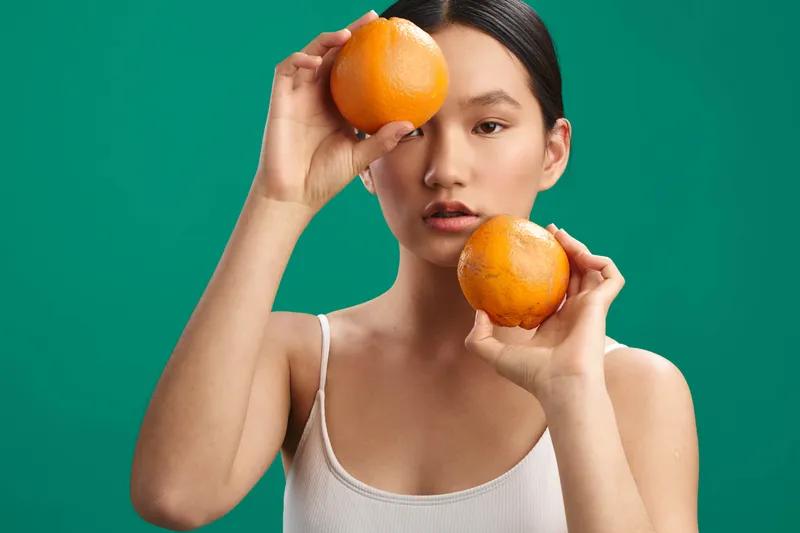asian woman with dark hair in a white tank top holds two oranges near her face against a green backdrop, expressing playful curiosity and fresh, vibrant energy.