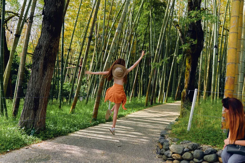 Woman in straw hat performs a joyful jump on a gravel path through bamboo forest, orange skirt flowing, carefree summer travel vibe, friend watching from stone bench while photographer captures