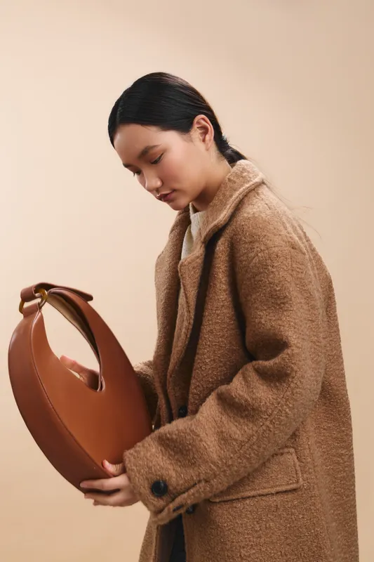 asian woman in a tailored coat examines a stylish circular handbag in a minimalist studio, conveying a thoughtful mood, fashion focus, and calm, modern style.