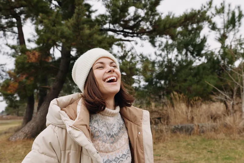 Woman in a warm coat and knit beanie laughs as she stands in a park with trees and dry grass, enjoying fresh air and sunshine on a chilly day, hair loose and friendly mood