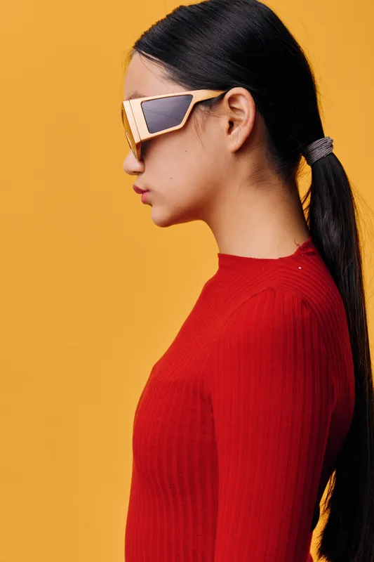 Woman with long black hair tied in ponytail wearing red sweater and futuristic sunglasses standing in profile on yellow background with minimal style and modern fashion look.