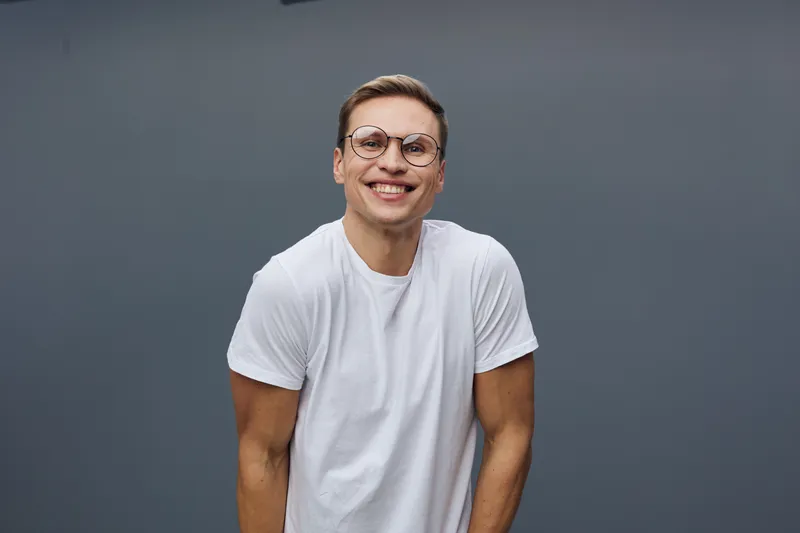 Smiling young man wearing glasses and white t-shirt looking at camera on plain gray background portrait. People lifestyle concept, positive expression, casual style.