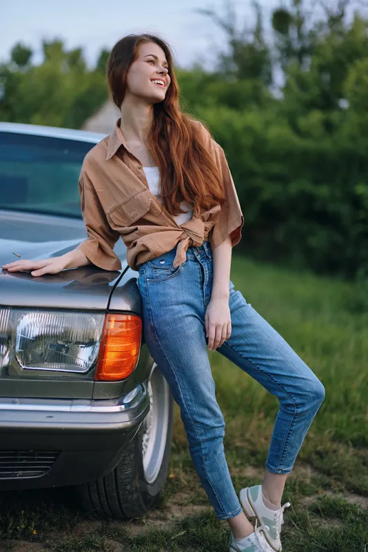 Young woman in casual attire smiling outdoors, leaning on vintage car, evoking feelings of joy and freedom in a lush green setting.