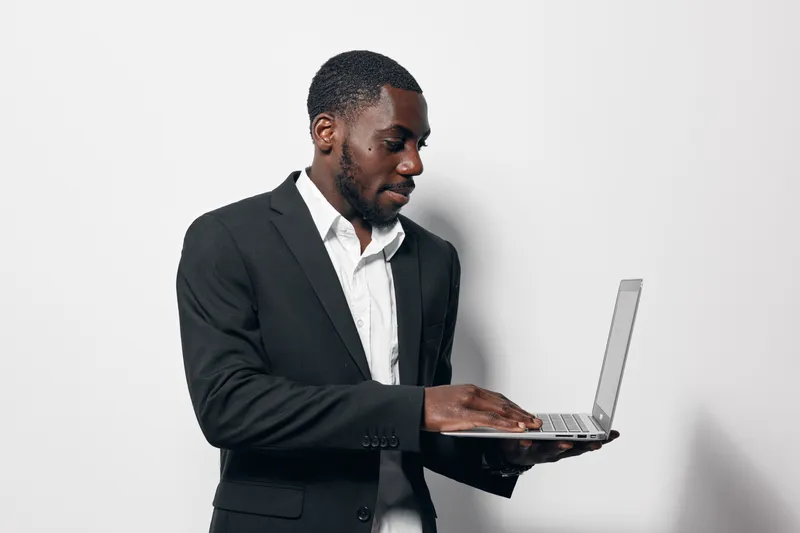 African American man in black suit using laptop computer for work or communication on white background. Business professional with dark skin focused on technology and office tasks.