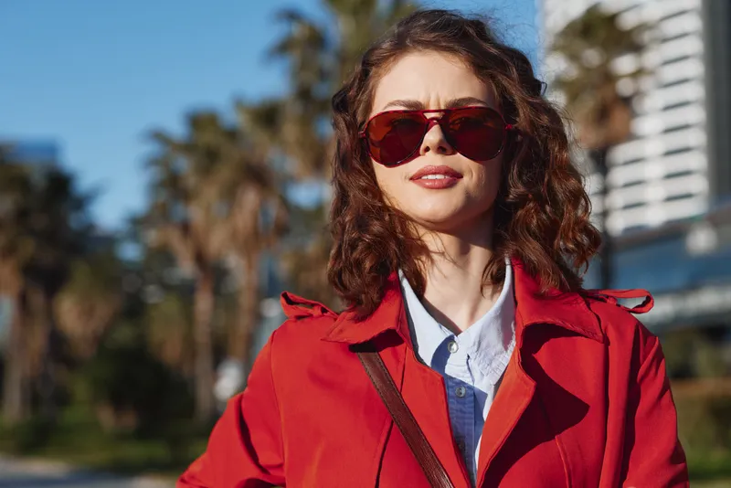 A stylish woman with curly brunette hair wears a red coat and sunglasses, standing outdoors on a sunny day, conveying urban confidence and fashion today for an editorial shoot
