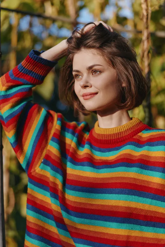 Woman portrait in colorful striped sweater smiling outdoors in autumn sunlight, short brown hair and relaxed pose with hand in hair, warm natural light and shallow depth.