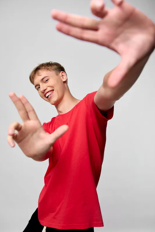 Hand gesture framing a smiling man in a red shirt, raised hands create a dynamic frame in a studio setting, capturing a playful, energetic and casual mood.