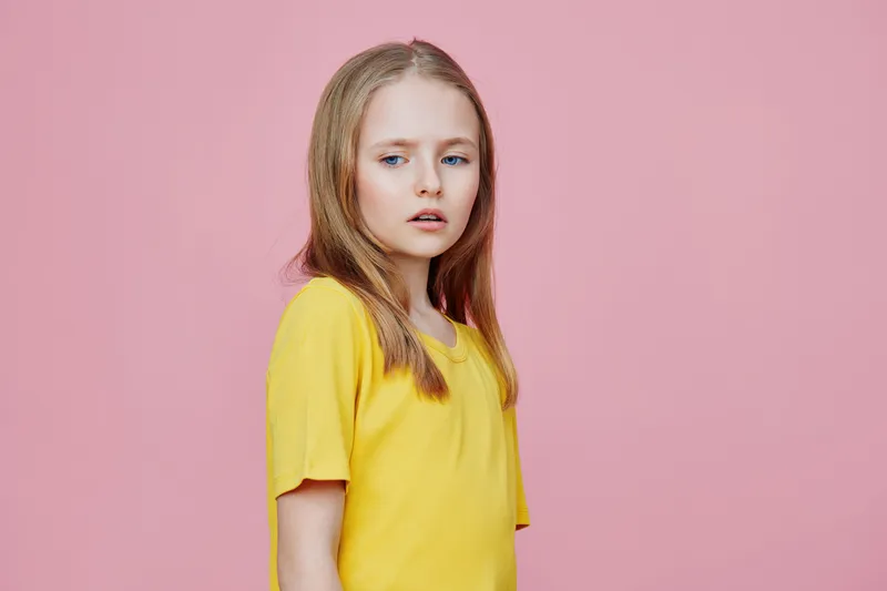 Young girl with long blonde hair wearing a yellow shirt against a pink background, expressing a thoughtful emotion, perfect for children related themes