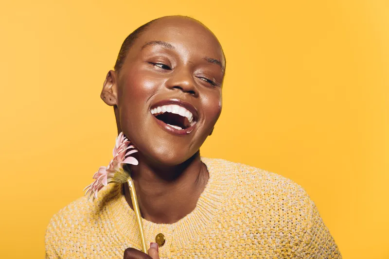 Smiling dark skinned African woman in yellow sweater holds pink flower near face on yellow background. Concept of happiness, beauty, natural skin, and joyful lifestyle portrait.