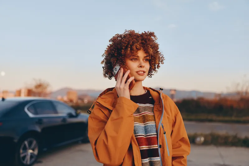 Smiling woman with curly hair speaking on smartphone outdoors at sunset near parked cars, orange jacket and casual lifestyle, warm city light, phone call communication with travel vibe