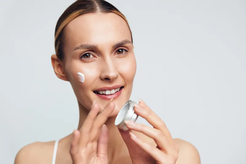 A smiling young woman applies white face cream on her cheek while holding a small open jar in a bright studio. Concept for skincare, beauty routine, and healthy skin care.