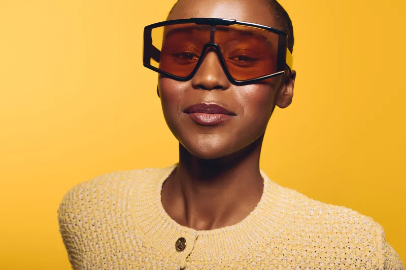 Confident African woman with dark skin wearing large sunglasses and a knitted yellow sweater posing on yellow background. Concept of fashion, beauty, and modern style portrait.