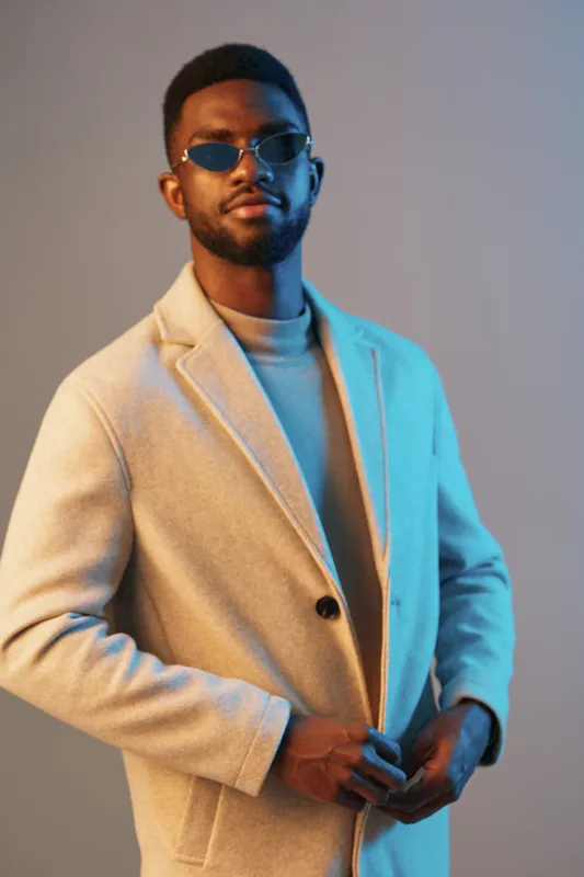 Stylish young man in a beige coat and sunglasses, showcasing a confident expression against a gradient blue background, ideal for fashion and seasonal promotions