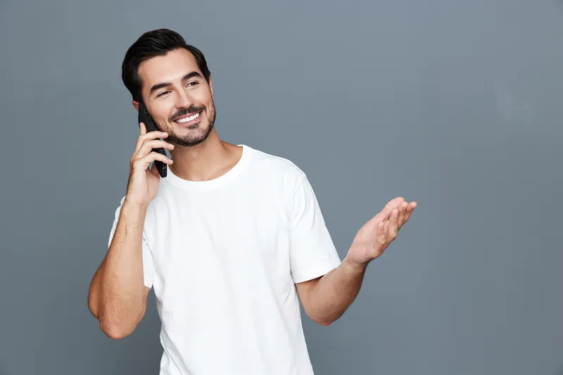 Smiling man wearing casual white t-shirt talking on mobile phone with joyful expression, isolated on plain solid gray background. Communication lifestyle concept.