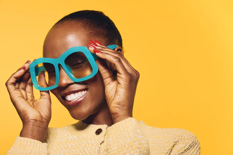 Smiling black woman with dark skin wearing large blue sunglasses and beige sweater poses on yellow background. Concept of fashion, beauty, summer style, and confident African ethnicity model.