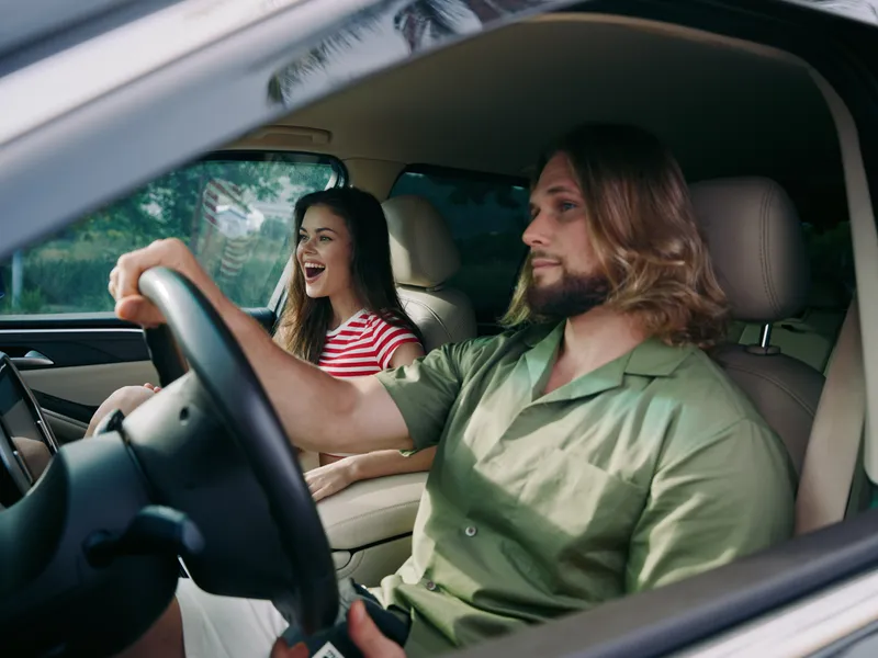 A couple sits inside a car with the driver smiling and the passenger laughing, captured in a bright, candid moment during a road trip.