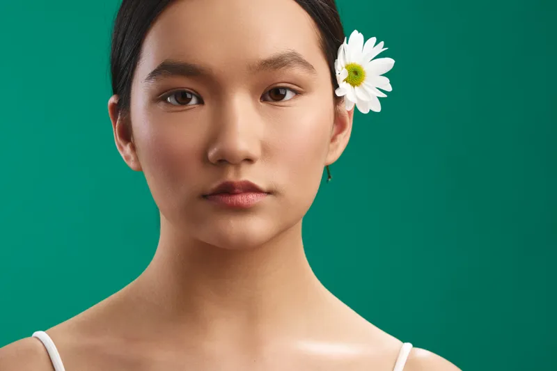 Asian woman with a calm, composed expression stands against a teal backdrop in a closeup portrait, accented by a white flower in her hair for a graceful, contemporary vibe.