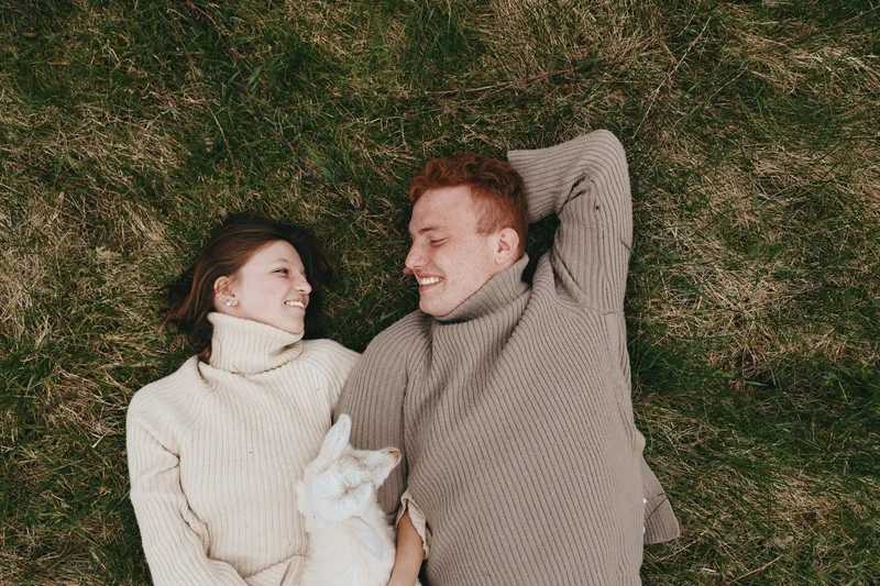 Cozy couple lying on grass outdoors, wearing matching knit sweaters, warm tones and soft light, relaxed smiles, intimate connection, carefree lifestyle moment in nature, gentle breeze