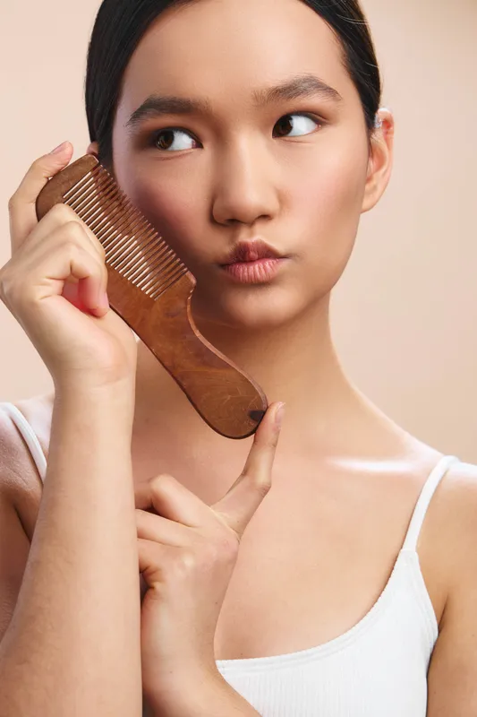 Asian woman holds a wooden comb to her hair in a soft studio setting, exuding calm and focus as she cares for her appearance.