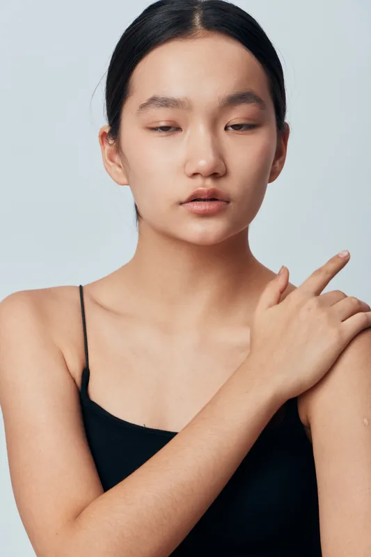 Young woman with clear skin and natural makeup touching shoulder wearing black sleeveless top against gray background. Portrait of calm female model with neutral expression in studio.